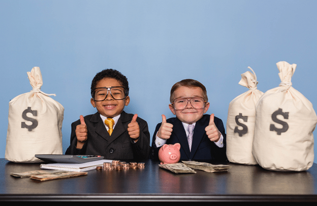 two boys sitting at desk dressed in suits as bankers surrounded by money bags, coins and paper money displaying thumbs up signs