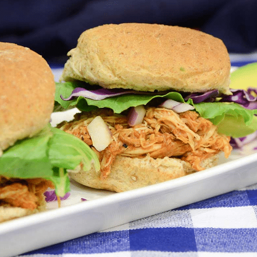 Pulled chicken sliders with red onion, red rabbage and greens on long white plate at blue gingham clad outdoor table