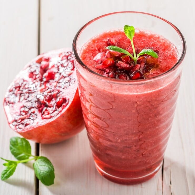 pink smoothie in glass cup with mint garnish next to a halved pomegranate